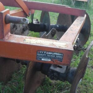 DSCF4522 - Montana Auctions by Kevin Hill Rusty orange tillage tool with metal discs on grass; label reads Pittsburgh Quality Tillage Tools.