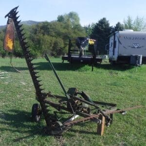 DSCF4531 - Montana Auctions by Kevin Hill Rusty spiked hay rake on a wheeled frame in a grassy field, with a trailer and camper in the background.