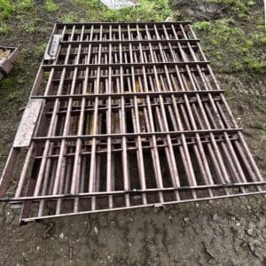 Rusty metal grate panel lying on muddy ground outdoors, with grass and scattered debris around it.