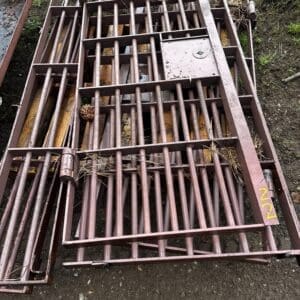 Pile of rusted metal gate frames with vertical bars stacked outdoors on dirt, with scattered hay and a pinecone nearby.