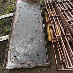 Long rusty steel trough with peeling paint and several drainage holes sits on dirt beside rusty metal frames and cages in an outdoor yard.