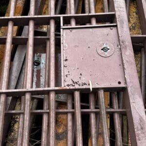 Rusty metal grate grid with an access panel featuring a small padlock, outdoors on weathered wood.