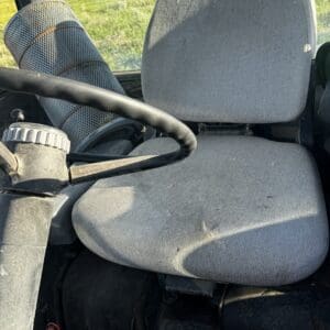 Grey fabric tractor seat and steering wheel inside a cab with a sunlit grassy field outside.”] ,