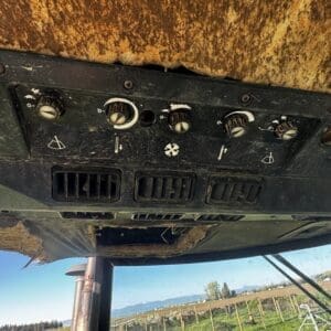 IMG_1307 - Montana Auctions by Kevin Hill Dusty control panel with knobs and vents inside a worn tractor cab; outside, a rural field and mountains are visible.