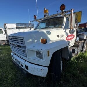 IMG_1309 - Montana Auctions by Kevin Hill Angled view of a white utility tow truck with a Pierce Homes logo, rusted equipment, and amber warning light on a grassy lot.