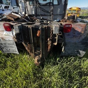 IMG_1311 - Montana Auctions by Kevin Hill Rear view of a truck with an 'Oversize Load' sign, tangled hoses and chains, rusted hitch, and tall grass in the foreground; a bus in the distance.