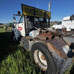 IMG_1313 - Montana Auctions by Kevin Hill White tow truck with a yellow 'OVERSIZE LOAD' sign, rusted bed, parked on grass under a clear blue sky.