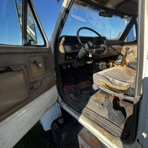 IMG_1314 - Montana Auctions by Kevin Hill Interior of an old, rusted truck cab with door open, dusty brown dashboard and steering wheel, and a worn seat with debris on the floor on a sunny day.