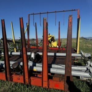 IMG_1320 - Montana Auctions by Kevin Hill Rusty red farm trailer with tall vertical stakes and scattered metal pipes on a grassy field under a blue sky.