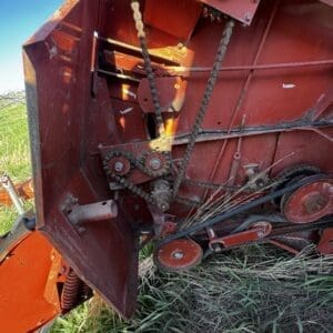 IMG_1329 - Montana Auctions by Kevin Hill Close-up of a red farming machine showing gears, chains, and belts with grass in a field behind it
