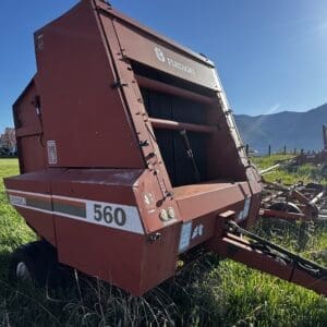 IMG_1333 - Montana Auctions by Kevin Hill Red agricultural baler labeled 560 parked in a grassy field with mountains in the distance; hardware and hitch visible in sunlight.
