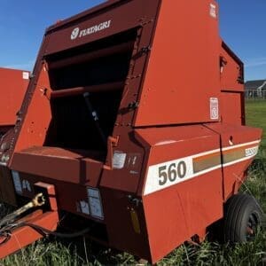 IMG_1334 - Montana Auctions by Kevin Hill Red Fiatagri 560 round baler viewed from the rear-left in a grassy field under a clear blue sky, showing the open intake and belt mechanism.