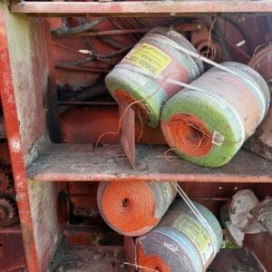 IMG_1337 - Montana Auctions by Kevin Hill Three orange wire rope spools stored in a dusty red metal toolbox on farm machinery, with coils and loose strands visible.