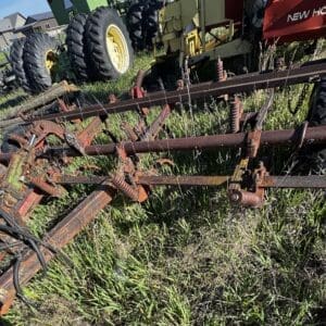 IMG_1342 - Montana Auctions by Kevin Hill Rusty agricultural implement with multiple bars and springs on a grassy lot; New Holland machinery visible in the background.