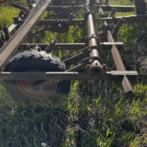 IMG_1344 - Montana Auctions by Kevin Hill Rusty farm implement (cultivator/harrow) on a metal frame with a wheel, resting in tall grass in a field of weeds.