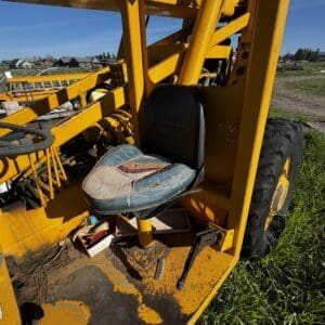 IMG_1349 - Montana Auctions by Kevin Hill Yellow construction vehicle frame with a cracked, torn seat, parked on a dirt-and-grass lot with other equipment in the background.
