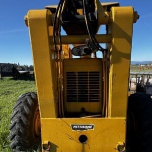IMG_1356 - Montana Auctions by Kevin Hill Rear view of a yellow Pettibone heavy equipment machine with large tires and exposed hydraulic hoses in a grassy field under a blue sky.