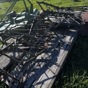 IMG_1361 - Montana Auctions by Kevin Hill Weathered wooden trailer bed with rusted agricultural harrow/cultivator frame lying on it in a sunlit grassy field, with a fence and buildings in the background.