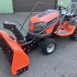 Red riding lawn mower with front-mounted orange snow blower attachment and tire chains on rear wheels.