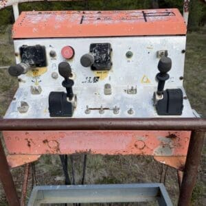 IMG_6543 - Montana Auctions by Kevin Hill Weathered orange control panel with two joysticks and levers outdoors, rusted frame, trees in background.