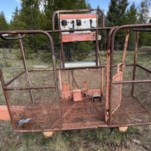 IMG_6544 - Montana Auctions by Kevin Hill Rusty metal aerial work platform basket with mesh sides and a control console, sitting in a grassy, wooded area.