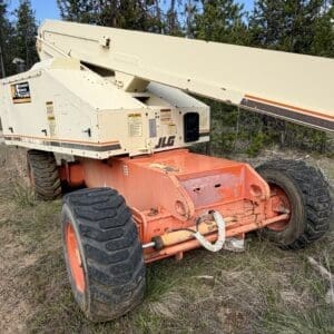 IMG_6545 - Montana Auctions by Kevin Hill Orange and cream mobile boom lift (JLG) on rugged ground with large tires, ready to lift operations near a forested area.