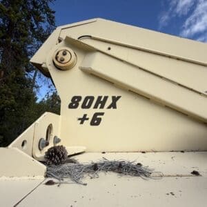 IMG_6546 - Montana Auctions by Kevin Hill Beige heavy equipment panel with model text '80HX +6' outdoors; nearby pine cone, needles, and tangled wire on the surface.