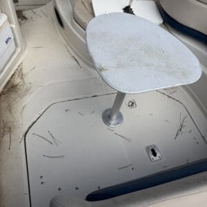 IMG_6554 - Montana Auctions by Kevin Hill Cockpit of a boat with a weathered rounded table on a pedestal, beige seating, and a deck scattered with pine needles.