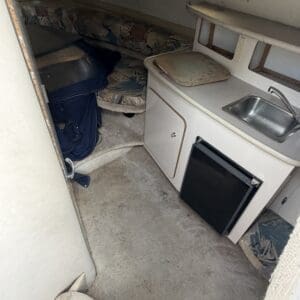 IMG_6557 - Montana Auctions by Kevin Hill Compact boat cabin featuring a small galley: sink, countertop, cutting board, and a mini fridge beneath; cushions and a bed area visible in the background.