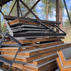 IMG_6575 - Montana Auctions by Kevin Hill Stack of rusted orange metal frames and assorted construction panels piled under a timber shelter with trees in the background.