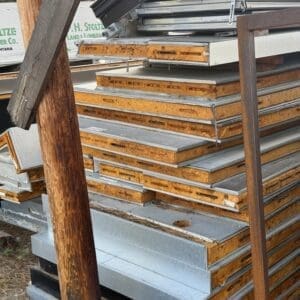 IMG_6577 - Montana Auctions by Kevin Hill Pile of rusted orange metal panels and galvanized sheets stacked outdoors, with a wooden post in the foreground on a construction site