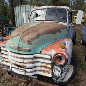 IMG_6578 - Montana Auctions by Kevin Hill Rusty vintage Chevrolet pickup with peeling teal and orange paint, cracked windshield, missing headlight, and heavy rust on the grille in a junkyard setting.