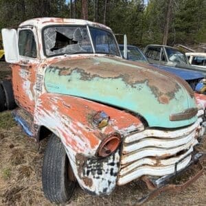 IMG_6580 - Montana Auctions by Kevin Hill Rusty vintage truck with peeling teal and orange paint in a grassy junkyard, cracked windshield and bent chrome grille faded by time.