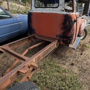 IMG_6581 - Montana Auctions by Kevin Hill Rear view of a weathered orange and white vintage pickup cab on a rusty trailer frame in a rural yard with trees nearby