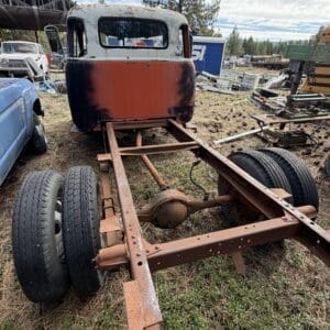 IMG_6583 - Montana Auctions by Kevin Hill Rusty truck chassis with a faded orange and white cab, missing bed, on a grassy junkyard lot.