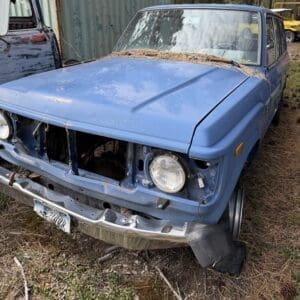 IMG_6588 - Montana Auctions by Kevin Hill Abandoned blue vintage car in a junkyard with a missing grille and rusted bumper, pine needles on the hood.