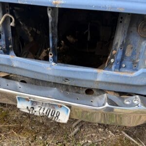 IMG_6589 - Montana Auctions by Kevin Hill Rusty blue vintage truck with its front grille and headlights removed, exposing a dark, empty engine bay and rusted metal frame. A weathered Arizona license plate hangs crookedly from the bumper. Ground covered in dry grass and weeds nearby.