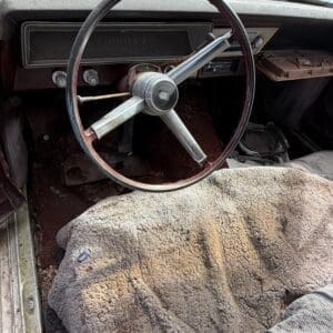 IMG_6605 - Montana Auctions by Kevin Hill Old car interior with a metal three-spoke steering wheel, dusty dashboard, and a worn shag carpet covering the front floor.