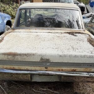 IMG_6607 - Montana Auctions by Kevin Hill Rear view of a dusty, rusted vintage car in a junkyard, chrome bumper visible and surrounding debris on the trunk area.