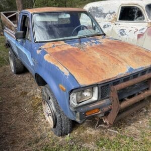 IMG_6609 - Montana Auctions by Kevin Hill Blue old pickup truck with a heavily rusted hood, missing its front wheel and a rusty bumper, parked in a grassy junkyard beside another weathered car.