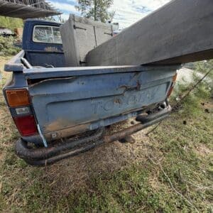 IMG_6612 - Montana Auctions by Kevin Hill Old blue Toyota pickup with a long wooden beam loaded in the bed, parked on grassy, overgrown ground; visible rust and a rundown rear bumper in a rural setting.