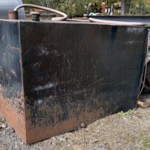 IMG_6615 - Montana Auctions by Kevin Hill Large black metal storage tank with rust along the bottom, outdoors on dirt, with a coil of hose on top and machinery visible at the right.
