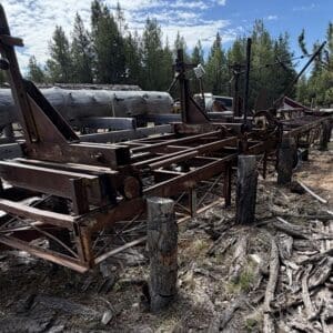 IMG_6618 - Montana Auctions by Kevin Hill Rusty vintage farming equipment laid out on a dirt lot beside stacked logs and pine trees in a forested yard.