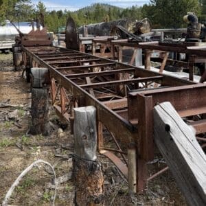 IMG_6624 - Montana Auctions by Kevin Hill Rusty metal framework and wooden posts on an outdoor, forested industrial site with logs and equipment in the background.