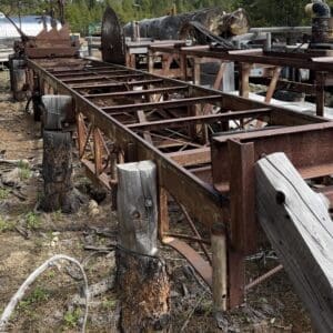 IMG_6625 - Montana Auctions by Kevin Hill Rusted metal framework and weathered wooden posts in an outdoor industrial yard with logs in the background.