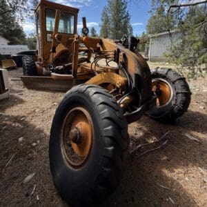 IMG_6635 - Montana Auctions by Kevin Hill Large orange construction vehicle (bulldozer/grader) with a front blade, exposed hydraulic arms, and oversized tires parked on a dirt lot amid trees and buildings.