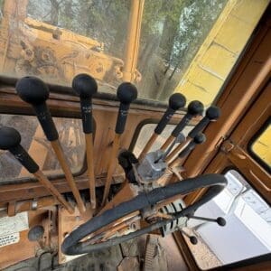 IMG_6638 - Montana Auctions by Kevin Hill Interior of a dusty orange excavator cab with multiple black-lever controls and a worn steering wheel seen from the operator's seat