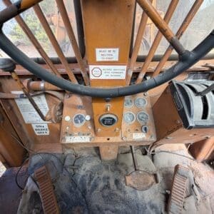 IMG_6640 - Montana Auctions by Kevin Hill Close-up of an orange construction vehicle cab: worn pedals, steering wheel, and a central control panel with gauges and switches; safety label reads 'Must be in neutral to start'