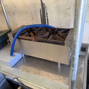 IMG_6642 - Montana Auctions by Kevin Hill Industrial stainless steel gas burner with four cast-iron grates and a blue hose resting on top.