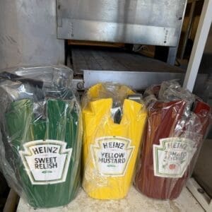IMG_6645 - Montana Auctions by Kevin Hill Three Heinz condiment bottles in plastic wrap: green Sweet Relish, yellow Mustard, and red Ketchup on a stainless counter near a grill.
